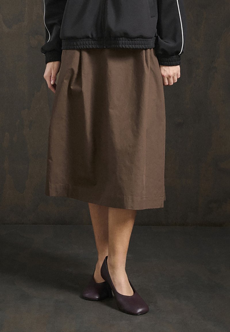 Pruun põlvikute pikkusega seelik, mis on valmistatud puuvillast materjalist, omades lihtsat disaini ja pehmet tekstuuri. Kombineeritud tumelilla kontsakingadega.