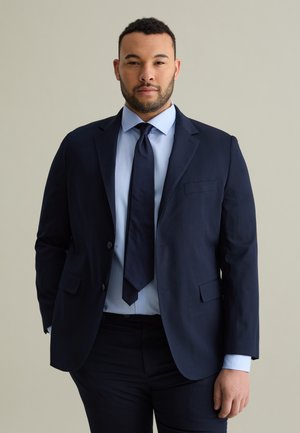 Navy suit jacket with a notched lapel, single chest pocket, paired with a light blue shirt and navy tie. Smooth fabric, tailored fit.