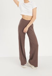 Bruna ribbade vida byxor med hög midja, tillsammans med en vit kortärmad t-shirt och ljusa sneakers, mot en enkel bakgrund.