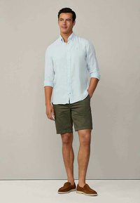 Camisa azul clara de botões, com as mangas enroladas, combinada com calções verdes-oliva. Sapatos castanhos tipo slip-on completam o visual casual.