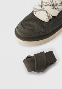 Μύτη αθλητικού παπουτσιού σκούρου πράσινου με λευκά κορδόνια με σχέδιο δεμένα γύρω από τον αστράγαλο και ένα επιπλέον ζευγάρι σκούρων πράσινων κορδονιών δίπλα του.