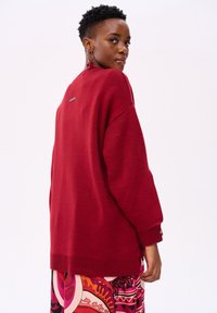Rode oversized trui met een ribbelboord aan de onderkant en mouwen, met een klein label aan de achterkant. Draag het over een kleurrijke, gemusterde rok.