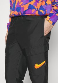 Černé cargo kalhoty s bočními kapsami, na kterých je oranžové logo Nike. V kombinaci s barevným svrškem s vzorem v purpurové, oranžové a modré barvě.
