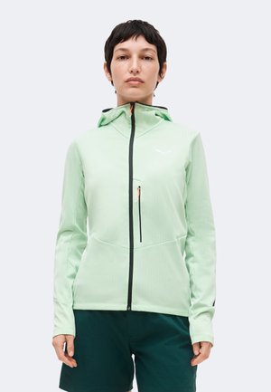 Žena stojaca čelom, oblečená v svetlozelenú mikinu na zips s náprsným vreckom a tmavozelené kraťasy na jednotnej farbe pozadia.