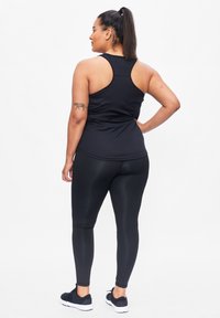 Zwarte sporttop met racerback ontwerp, gecombineerd met glanzende zwarte leggings. Model draagt zwarte sneakers en toont een tatoeage op de linkerarm.