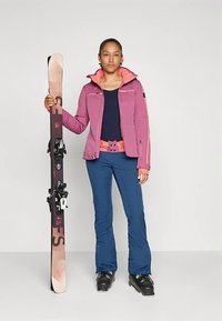 Echipament de schi format dintr-o geacă roz cu accente portocalii, pantaloni bleumarin și bocanci de schi negri, ținând o schiță cu legături.