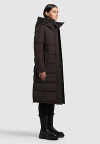 Braune Daunenjacke mit hohem Kragen, gestepptem Design, Reißverschluss vorne und Seitentaschen. Getragen mit schwarzen Leggings und klobigen schwarzen Stiefeletten.