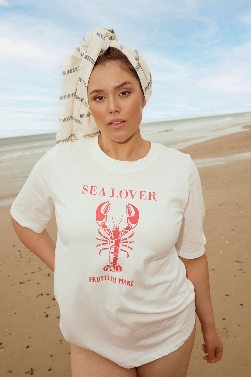 Femme sur une plage de sable portant un t-shirt blanc « SEA LOVER » avec un graphique de homard rouge et une serviette enroulée autour de sa tête sous un ciel partiellement nuageux.