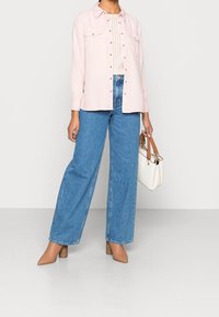 Lichtroze overhemd met knopen en twee borstzakken, gecombineerd met hooggetailleerde blauwe wide-leg jeans en beige schoenen met hak. Witte handtas met bruine bandjes.