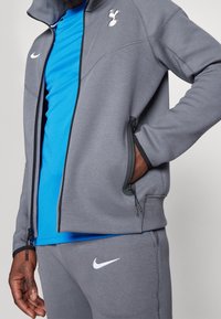 Giacca atletica grigia con tasche con zip e dettagli neri, caratterizzata da un logo dei Spurs. Indossata sopra una camicia blu, che mette in evidenza un design sportivo.