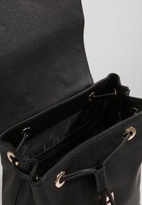 Bolso tipo bucket negro hecho de material sintético texturizado, con un borde ajustado en la parte superior, ojales en tono dorado y múltiples compartimentos interiores.