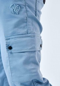 Pantalones cargo azules de tela ligera que cuentan con un bolsillo lateral con un botón negro y un logotipo bordado en la parte superior de la pierna.