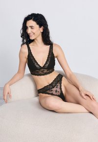 Soutien em renda preta com bordas recortadas e padrões florais, combinado com tanga em renda a condizer, ambos com painéis transparentes.