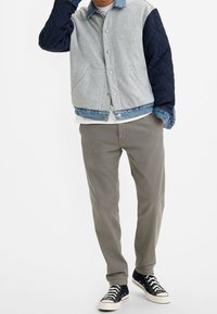Veste matelassée grise et bleu marine avec des accents en denim, associée à un pantalon gris clair et des baskets montantes noires avec des lacets blancs.