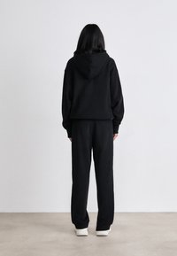 Melnas kapuces džemperis un atbilstošas sporta bikses. Audums izskatās mīksts; satur lielu kapuci un brīvu piegriezumu. Tenisēm ir gaiša krāsa.