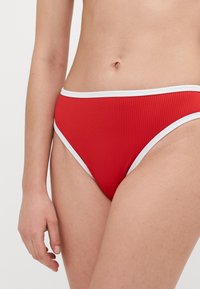 Vörös bordázott bikini alsó fehér díszítéssel, magas kivágású lábrészekkel és rugalmas derékkal. Sima textúra, finom fényességgel.