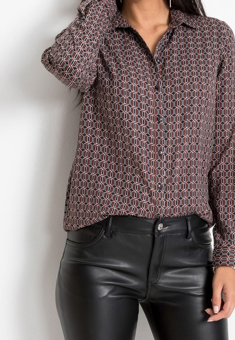 Femme portant une chemise à manches longues à motifs boutonnée, rentrée dans un pantalon en cuir noir taille haute, debout contre un mur uni.
