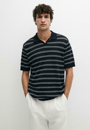 Jonge man met krullend haar die een zwart poloshirt met witte horizontale strepen en een witte broek draagt, staand met handen in de zakken.