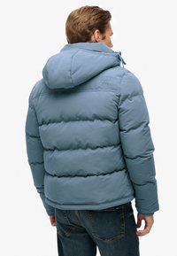 Chaqueta de abrigo azul con capucha, con acolchado de colcha, costuras en contraste y un pequeño logo en la parte superior de la espalda. Llevada con jeans oscuros.