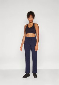 Žena stojaca proti jednoduchému pozadi, oblečená v čiernom športovom nátelníku, námorníckych teplákoch s šnúrkou a čiernych športových topánkach.