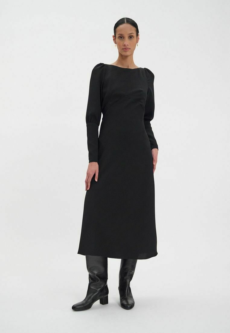 Robe noire à manches longues avec un décolleté carré, corsage ajusté et jupe fluide. Le modèle porte des bottes noires jusqu'au genou. Texture de tissu lisse.