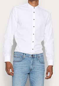 Valkoinen pitkähihaine paita, jossa on pystykraava ja mustat napit edessä, yhdistettynä vaaleansinisiin farkkuihin.