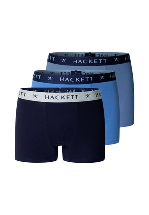 Jonge volwassen man staat blootsvoets tegen een grijze muur, draagt blauwe boxershort met een zwarte tailleband met label "HACKETT."
