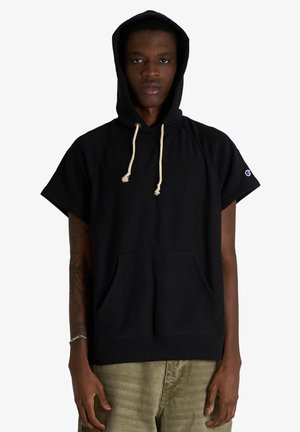 Uomo che indossa una felpa nera con cappuccio a maniche corte con coulisse e tasca frontale, abbinata a pantaloni marrone chiaro su uno sfondo semplice.