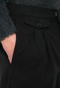 Μαύρο παντελόνι από υφή ύφασμα, με πλισέ σχεδιασμό, πλαϊνή τσέπη και κλείσιμο με μαύρο κουμπί. Συνδυάζεται με ένα χνουδωτό γκρι πουλόβερ.