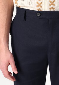 Navyblauwe broeken met een gladde textuur, voorzien van een enkele knoopsluiting en zijzakken. Pasvorm op maat met een subtiel diagonale weefpatroon.