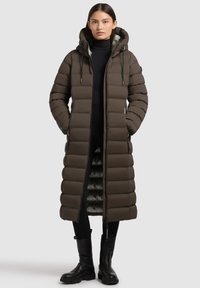 Giacca lunga imbottita marrone con cappuccio, caratterizzata da sezioni trapuntate, cerniere e una fodera testurizzata. Indossata sopra abiti neri e stivali.