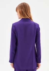 Blazer viola con colletto e maniche lunghe, caratterizzato da un tessuto testurizzato e un taglio dritto. La vista posteriore mostra linee fluide e dettagli minimi.