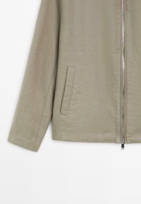 Manche de veste beige avec une fermeture éclair verticale à l'avant et une seule poche latérale rectangulaire sur le côté gauche.