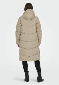 Abrigo largo beige acolchado con capucha, con líneas de acolchado horizontales y corte recto, combinado con pantalones negros y botas de tacón.