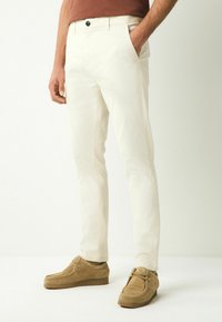 Pantalones de algodón blancos con un ajuste slim, que cuentan con bolsillos frontales y un botón negro. Combinados con zapatos de ante marrón claro.