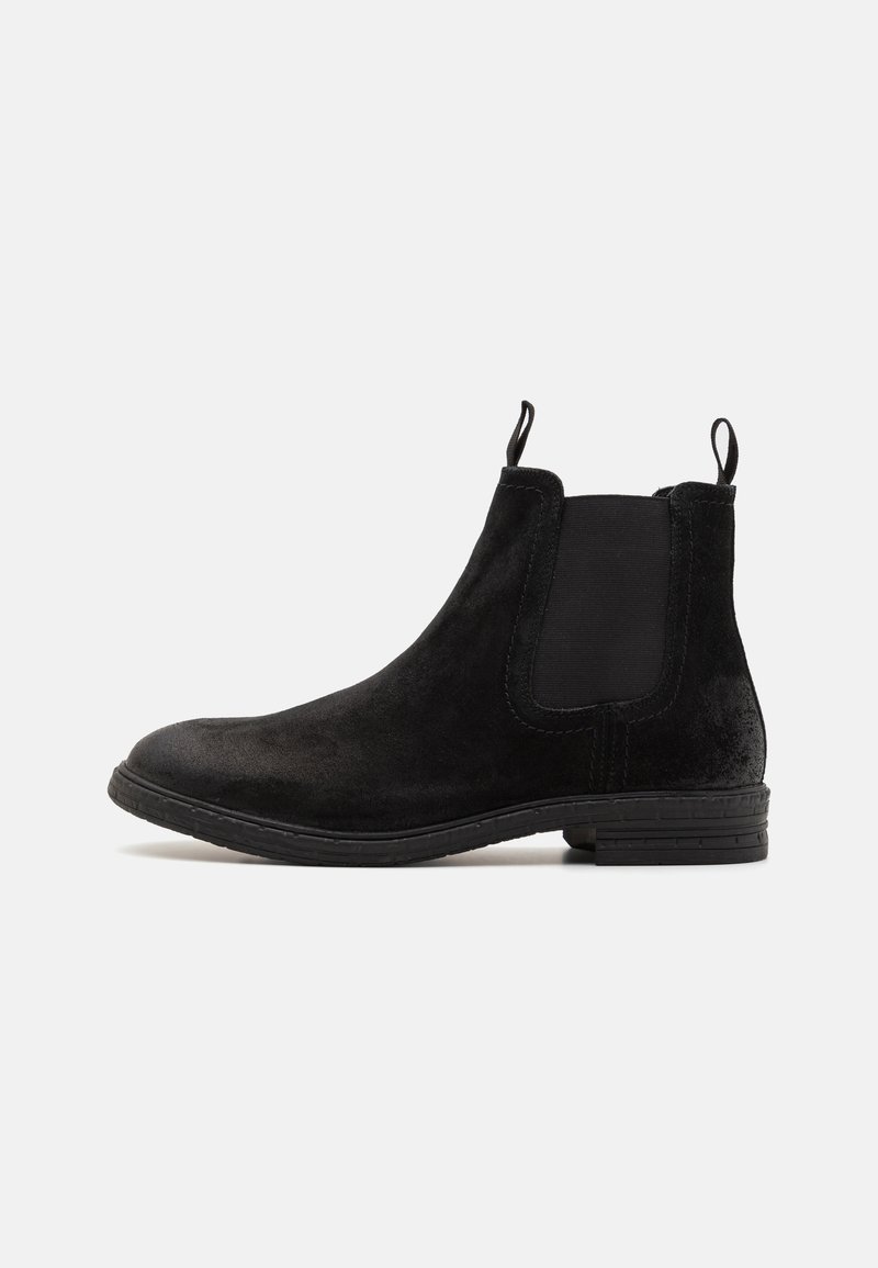 Bottine en daim noire avec panneaux latéraux élastiques, languettes à l’avant et à l’arrière, et un petit talon empilé sur fond blanc.