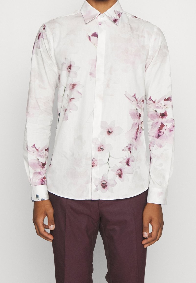 Chemise blanche à manches longues avec boutonnière, imprimé floral rose clair et violet, portée avec un pantalon violet foncé, vue du cou jusqu'aux hanches.