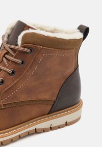 Brun læderstøvle med en shearling-foring, metaløjer, kontrasterende brune detaljer og en robust gummisål med markant mønster.