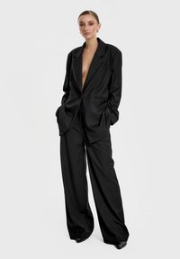 Czarna marynarka dopasowana o głębokim dekolcie w kształcie litery V i szerokich klapach, zestawiona z spodniami o szerokich nogawkach. Gładki materiał i zorganizowany fason.