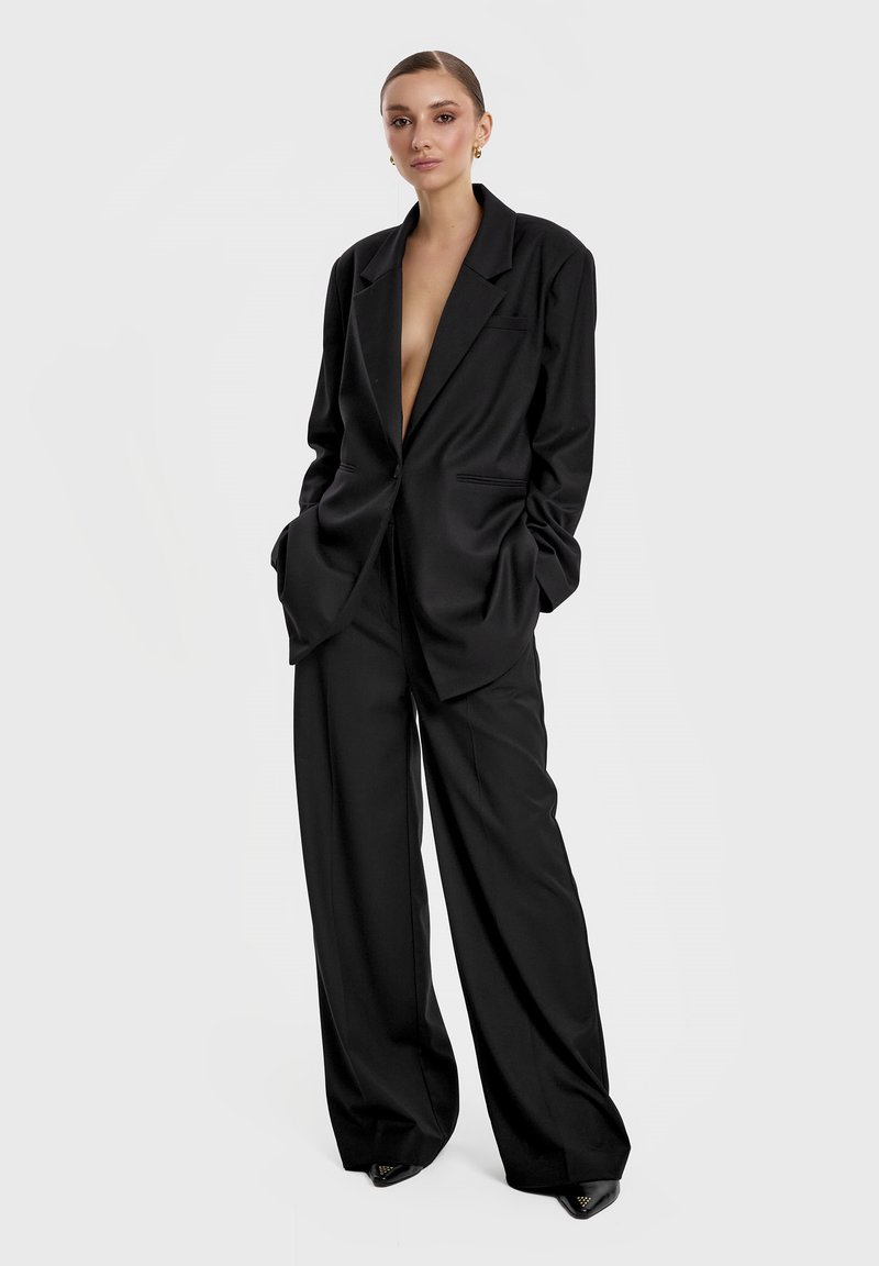 Czarna marynarka dopasowana o głębokim dekolcie w kształcie litery V i szerokich klapach, zestawiona z spodniami o szerokich nogawkach. Gładki materiał i zorganizowany fason.