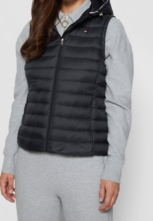 Sort puffervest med lynlås foran, prydet med vandret syninger, høj krave og hardware i guldfarve. Båret over en grå bluse med lange ærmer.