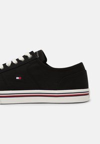 Schwarze Leinenschuhe mit weißen Schnürsenkeln, einer weißen Gummisohle und einem horizontalen roten und blauen Streifen. Beinhaltet ein kleines Logo an der Seite.