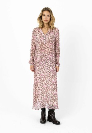 Frau mit blondem Haar, die ein langärmeliges Kleid mit pink-braunem Muster sowie schwarze Stiefel bis zur Mitte der Wade trägt und vor einem weißen Hintergrund steht.