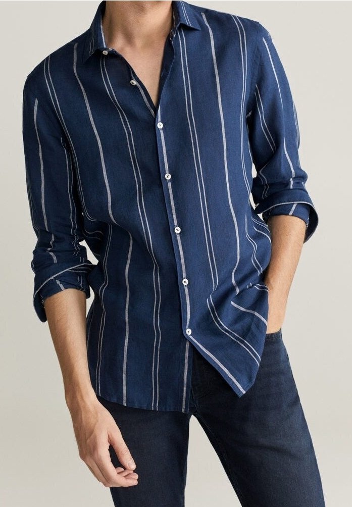 Homme portant une chemise bleu marine à boutons avec des rayures blanches verticales et un jean foncé, une main dans la poche, manches retroussées.