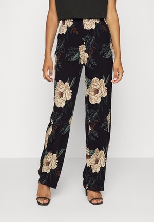 Pantalon noir à jambes larges avec un grand imprimé floral beige et vert, porté avec des sandales noires à talons hauts et fines lanières.