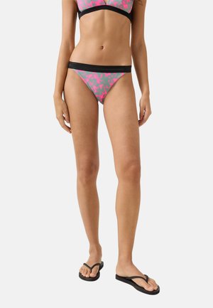 Frau trägt pink-grau gemusterten Bikini mit schwarzem Besatz und schwarzen Flip-Flops, steht vor einem schlichten weißen Hintergrund.