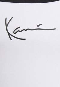 Biała odzież z czarnym nadrukowanym wzorem sygnatury; gładka faktura z kontrastowym czarnym dekoltem.