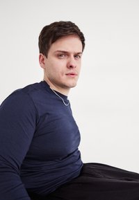 Tengerészeti hosszúujjú póló, testhezálló fazon, sima textúra. Ezüst lánc nyaklánc. Az alany ül, világos háttér, semleges kifejezés.