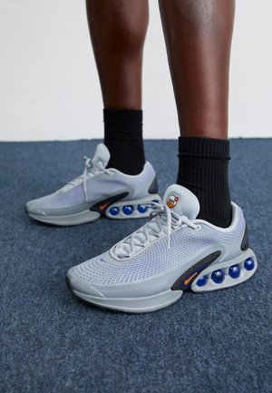 Jambes portant des chaussettes noires et des baskets Nike grises avec un amorti bleu sur un sol recouvert d'un tapis bleu, devant un mur blanc.