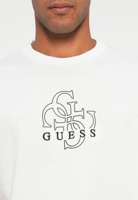 Biała bawełniana koszulka z krótkim rękawem z czarnym graficznym logo z przeplatającymi się literami "G" i "S", umieszczona na środku klatki piersiowej, z okrągłym dekoltem.